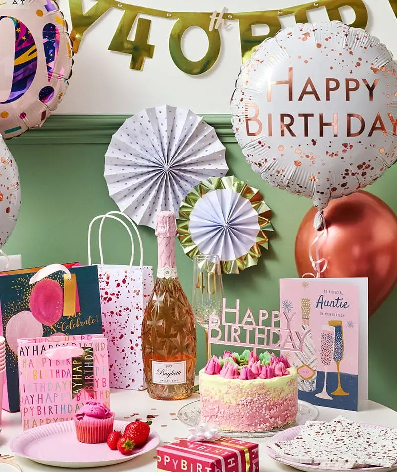  A colourful birthday party setup featuring balloons, gift bags, a pink cake with candles, a bottle of sparkling drink, greeting cards, and wrapped presents arranged on a decorated table.